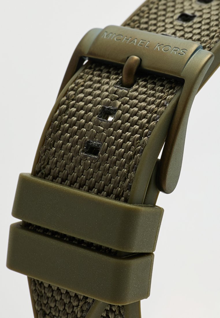 Primer plano de una correa de reloj de tejido verde oliva con agujeros rectangulares y hebilla de metal grabada "MICHAEL KORS."
