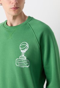 Grön sweatshirt med en vit trofégraphic och texten "Lower Manhattan Championship Tour 11 mars 1990" på bröstet.