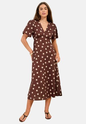 Vrouw draagt een bruine midi-jurk met beige polkadots, korte vleermuismouwen, V-hals en zwarte sandalen, staand met één hand in de zak.