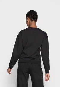 Sudadera negra de algodón con mangas largas, puños acanalados y cuello redondo. Textura lisa, sin estampados. Se lleva con pantalones negros, vista desde atrás.