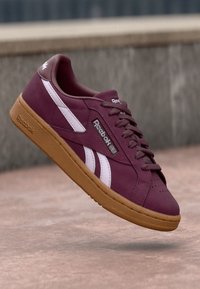 Burgunderfarbene Sportschuh mit weißen Streifen, aus Wildleder gefertigt, mit einer strukturierten Gummisohle und Metallösen für die Schnürsenkel. Sichtbares Reebok-Logo.