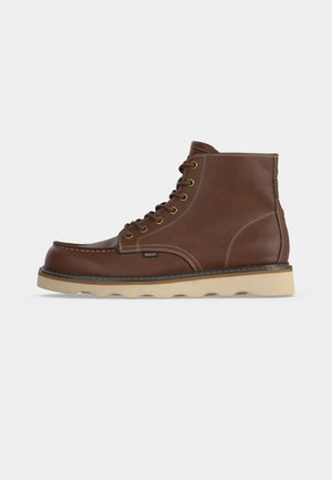 Bruine leren enkellaars met beige rubberen zool, veters aan de voorkant, zichtbare stiksels en een klein label aan de zijkant.