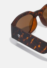 Tortoiseshell sunglasses with brown lenses, wide square frame, and textured arms featuring a logo on the side.