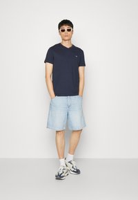 Camiseta de cuello en V color navy combinada con pantalones cortos de mezclilla azul claro. Zapatillas deportivas blancas con detalles en navy y calcetines blancos. Gafas de sol cuadradas en un tono claro.
