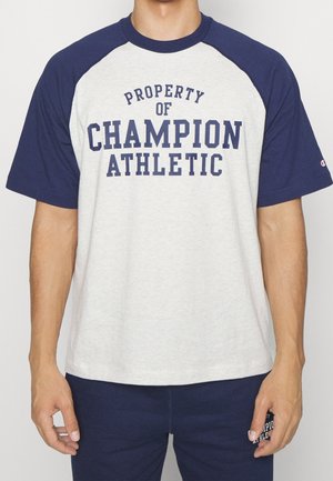 Person trägt ein weißes und marineblaues Raglan-T-Shirt mit der Aufschrift "PROPERTY OF CHAMPION ATHLETIC" vorne in marineblauen Buchstaben.