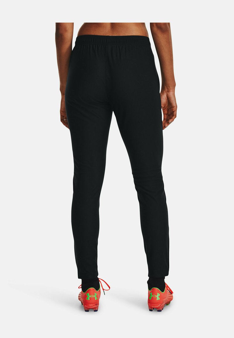 Pantalons de sport noirs avec une taille élastique, coupe ajustée et texture lisse, associés à des chaussures de football rouges vives avec des accents verts.