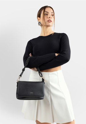 Donna che indossa un crop top nero a maniche lunghe e una gonna bianca, che tiene in mano una borsa nera in pelle testurizzata con tracolla intrecciata.