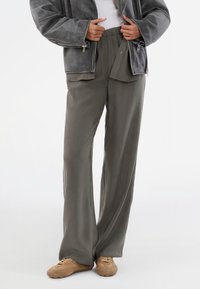 Veste en suède oversized grise portée sur un haut blanc et un pantalon léger gris clair ample. Des chaussures brunes complètent la tenue, mettant en avant un style décontracté.