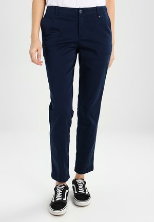 Persoon die marineblauwe slim-fit broek en zwart-witte veter-sneakers draagt, staand tegen een effen lichte achtergrond.