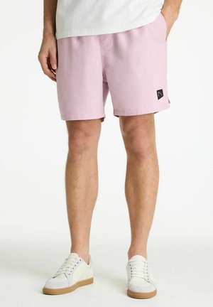 Bărbat purtând pantaloni scurți roz deschis și adidași albi, stând cu mâinile în buzunare pe un fundal alb simplu.