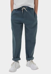 Osoba v modrozelených kalhotách s ohrnutými nohavicemi, bílým šňůrkovým páskem, bílých teniskách a šedých ponožkách na jednobarevném pozadí.