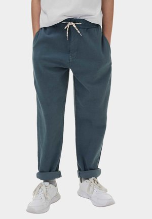 Person in teal Hochwasserhosen mit weißem Kordelzug, weißen Turnschuhen und grauen Socken vor einem einfarbigen Hintergrund.