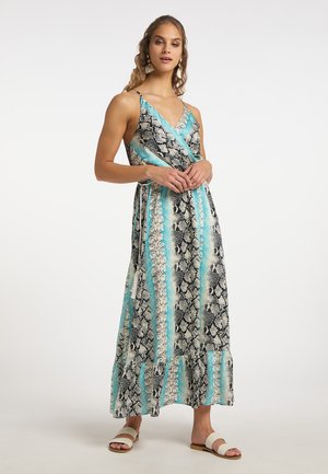 Vrouw draagt een mouwloze wikkeljurk met blauwe, zwarte en witte slangprint, met een rucheszoom en witte sandalen, staand tegen een effen achtergrond.