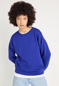 Felpa blu con vestibilità ampia, collo rotondo e spalle scese, indossata sopra una camicia bianca a strati, abbinata a jeans blu chiaro.
