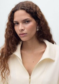 Veste zippée beige clair avec un col haut, présentant une texture lisse et un détail de fermeture éclair dorée visible. Cheveux longs et ondulés de couleur brune.