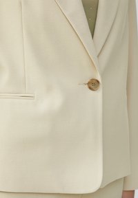 Blazer beige en tissu texturé, fermeture à un bouton et revers classique. Il présente une poche avant et une coupe ajustée.