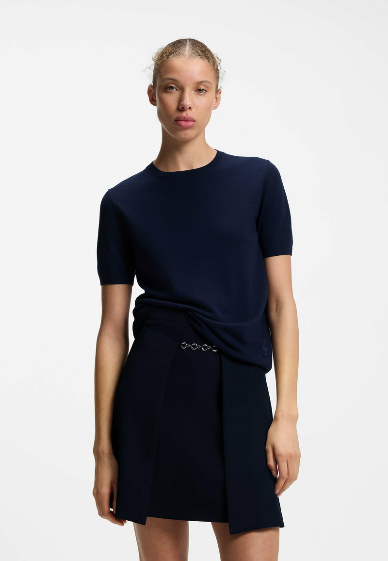Femme aux cheveux tirés en arrière portant un haut bleu marine à manches courtes et une jupe assortie, debout devant un fond blanc uni.