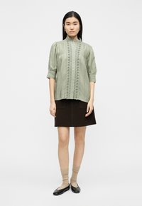 Γυναίκα που στέκεται φορώντας μπλούζα χρώματος φασκόμηλου με δαντέλα, σκούρο καφέ φούστα, μπεζ κάλτσες και μαύρες μπαλαρίνες, σε λευκό απλό φόντο.