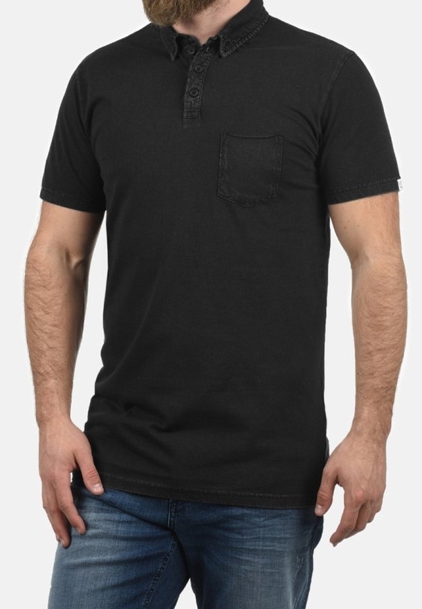 Homme portant un polo noir à manches courtes avec une poche poitrine et un jean bleu, debout, les mains détendues le long du corps.