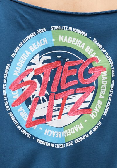 Μπλε ύφασμα με κυκλικό σχέδιο που περιλαμβάνει το «Stieglitz» με κόκκινα γράμματα, το «Madeira Beach», φοίνικες, πουλιά και «Island of Flowers 2026».