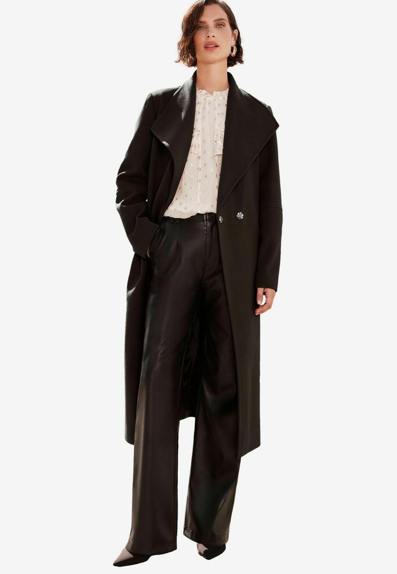 Svart ullblandad kappa med bred krage, dubbelknäppt stängning och långa ärmar; kombinerad med svarta högmidjade läderbyxor.