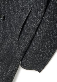 Giacca in lana grigio scuro con motivo a spina di pesce, caratterizzata da un bottone prominente, rever e dettagli sulle tasche laterali. Tessuto strutturato.