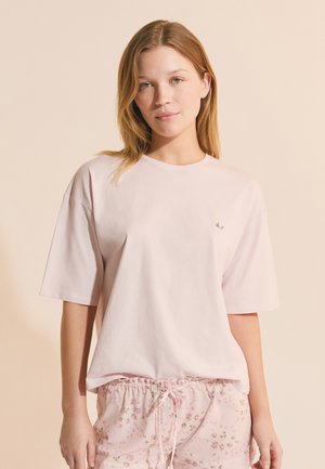 Mujer joven con cabello lacio de color castaño claro, viste una camiseta suelta de color rosa pálido y pantalones cortos con cordón a juego y estampado floral, de pie contra un fondo beige.