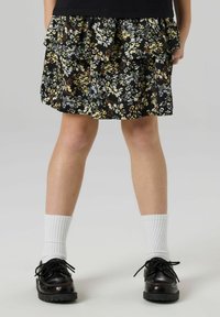 Melna ziedu raksta svārki ar bārkstainām kārtām, aizpildīti ar zaļām, brūnām un dzeltenām faktūrām; valkāti ar melnām kurpēm un baltām zeķēm.