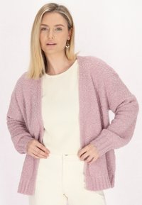 Teksturisan vaaleanpunainen neule, jossa on löysä istuvuus, avoin etuosa ja ribatut hihansuuhteet. Täydennetty vaaleancremellä paidalla ja valkoisilla housuilla.