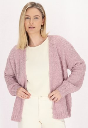 Cardigan rosa a trama con un fit morbido, apertura frontale e polsini a coste. Indossato sopra un top color crema, con pantaloni bianchi.