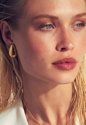 Guldiga droppörhängen, slät yta, polerad finish. Modellen har blött, rufsigt hår, neutral läppfärg och diskret ögonmakeup.