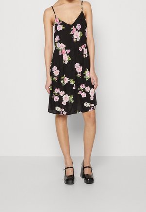 Mujer vestida con vestido floral sin mangas negro con flores rosas y blancas, combinada con zapatos Mary Jane negros de tacón alto, de pie sobre un fondo blanco.