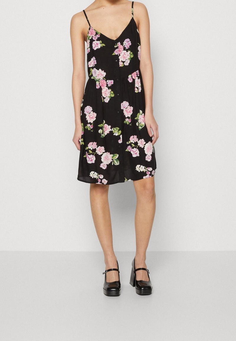 Femme portant une robe noire sans manches à fleurs roses et blanches, associée à des chaussures Mary Jane noires à talons hauts, debout devant un fond blanc.