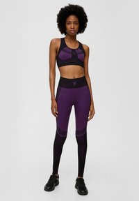 Lila Sport-BH mit Mesh-Akzenten und schwarzen Details, kombiniert mit hoch taillierten lila und schwarzen Leggings mit geometrischem Muster.
