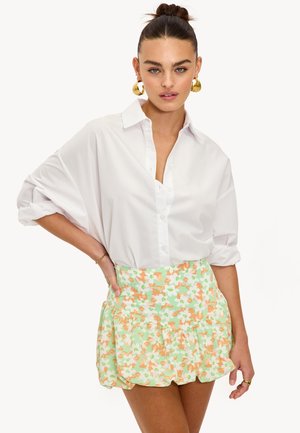 Chemise blanche oversize aux manches retroussées associée à une jupe verte à volants avec un motif floral orange et blanc.