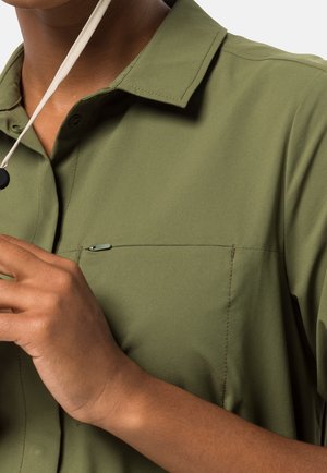 Person wearing an olive green shirt with a collar and chest pocket, adjusting a white cord necklace.