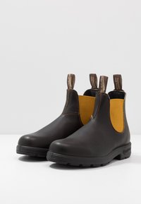 Botas de tornozelo em couro castanho com painéis laterais elásticos amarelos, biqueira arredondada, alças de puxar e sola de borracha texturizada. Detalhes de costura em contraste.