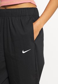 Svarta, lätta träningsbyxor med elastisk midja, sidofickor och en vit Nike-swoosh-logotyp på vänster lår.