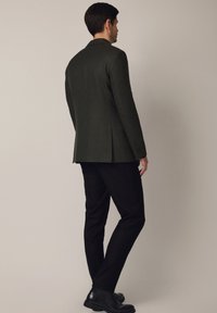 Ciemnozielony żakiet wykonany z teksturowanej tkaniny, z klasycznym kołnierzem, pojedynczym tylnym wentylatorem i dopasowanym krojem, w połączeniu z czarnymi spodniami.