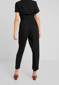 Jumpsuit noir à manches courtes, taille cintrée et jambes fuselées. Le matériau est lisse, avec un design épuré et minimaliste sans motifs visibles.