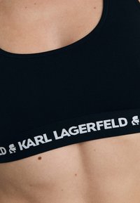 Top corto nero realizzato in tessuto elasticizzato con un ampio elastico che presenta il logo "Karl Lagerfeld" in bianco. Texture liscia e design semplice.