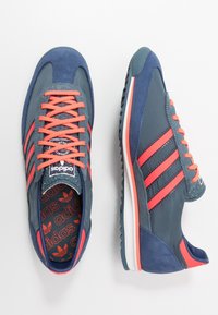 Granatowo-szare sneakersy z koralowymi sznurowadłami, trzema koralowymi paskami i teksturowanymi akcentami. Charakteryzują się miękką zamszową fakturą i białą podeszwą.