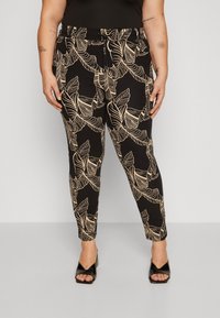 Kvinna som bär svarta skräddarsydda byxor med beige tropisk bladmönster och svarta sandaletter, som står mot en enfärgad grå bakgrund.