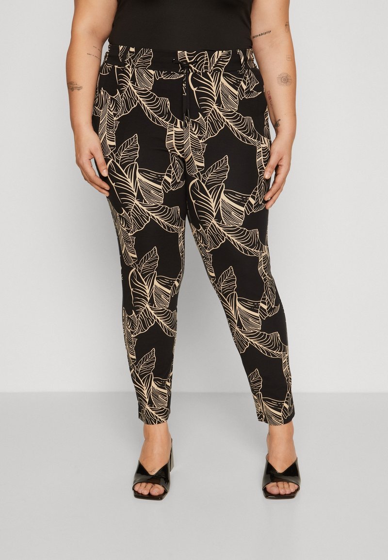 Kvinna som bär svarta skräddarsydda byxor med beige tropisk bladmönster och svarta sandaletter, som står mot en enfärgad grå bakgrund.