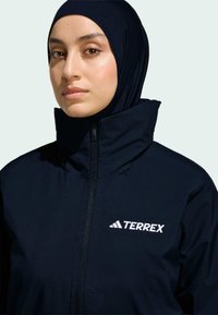 Námořnický Terrex kabát z látky s vysokým golierom, zipsom a bielym logom na hrudi. Hladká textúra, priliehavý strih a odolný materiál.