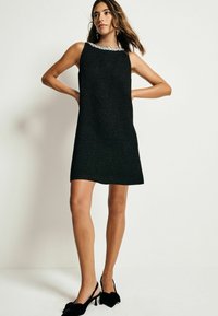 Robe noire sans manches avec un tissu texturé et un détail de col en perles. Paire de escarpins noirs à bride arrière avec des accents en noeud.