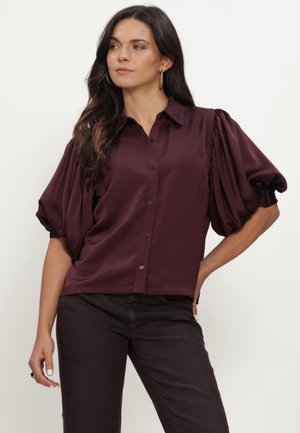 Vrouw die een donkerpaarse satijnen blouse met pofmouwen en een donkere broek draagt, staand met één hand op haar heup.