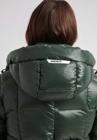 Grønne puffjakke med en skinnende, glat tekstur, stor hætte og syede sektioner. Har et hvidt logo-mærke på bagsiden.