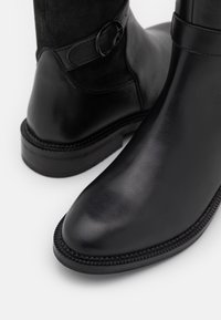 Bottes noires en cuir jusqu'à la cheville avec une finition lisse, une sangle en cuir et un détail en anneau métallique rond, semelle en caoutchouc texturé et bordure cousue.