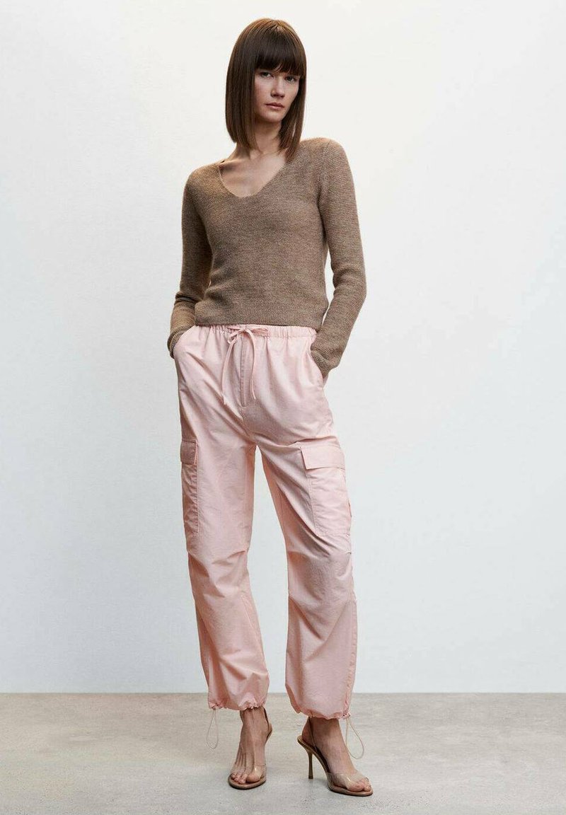 Suéter de punto marrón con escote en V, combinado con pantalones cargo rosa claro con tobillos elásticos y bolsillos laterales. Tacones transparentes completan el look.